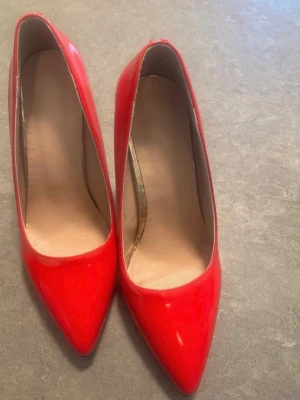 Röda lackpumps med spetsig tå - Säljer ett par klassiska röda pumps i glansigt lackmaterial med spetsig tå och smal klack. Insidan är beige och de har en elegant, tidlös design som passar till många olika stilar. Perfekta för dig som vill sticka ut med färg.