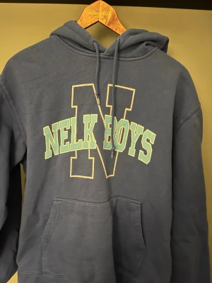 Blå Nelk Boys hoodie från Full Send - Snygg blå hoodie från Full Send med stor 'Nelk Boys'-logga i ljus blå text som har lite revor i texten där av priset 