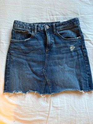 Blå jeanskjol med rå kant - Säljer en blå jeanskjol med rå, fransig kant och slitna detaljer framtill. Kjolen har klassisk femficksdesign, knapp och dragkedja i midjan. Perfekt för dig som gillar en avslappnad och trendig stil.