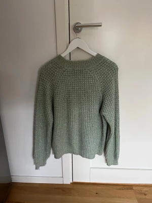 Ljusgrön stickad tröja - Mysig ljusgrön stickad tröja med grov struktur och rund hals. Tröjan har långa ärmar och ribbade muddar vid ärmslut och nederkant. Perfekt för kyliga dagar och enkel att matcha med jeans eller kjol.