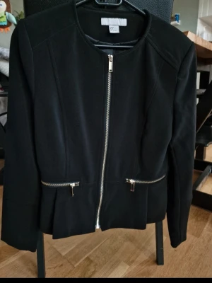 Svart kavaj med dragkedja H&M - Stilren svart kavaj från H&M med rundad krage och silverfärgad dragkedja framtill. Två fickor med dragkedjor ger en modern touch. Figurnära passform och lätt material gör den enkel att bära till olika looks. Knappt använd