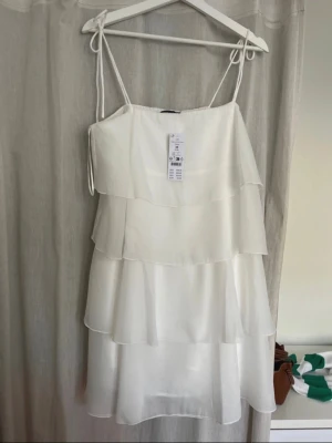 Vit volangklänning från Gina Tricot - Säljer en superfin vit klänning från Gina Tricot med tunna axelband och flera lager volanger. Klänningen är i ett lätt och luftigt material som ger en flowy känsla. Helt Ny! 