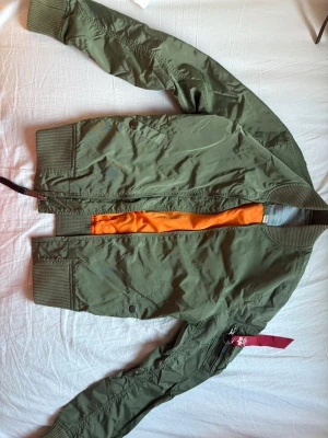 Grön bomberjacka från Alpha Industries - Klassisk grön bomberjacka från Alpha Industries med orange innerfoder och ribbade muddar. Jackan har dragkedja, fickor med tryckknapp och ikonisk röd tag på ärmen. Tillverkad i slitstarkt nylonmaterial och har en tidlös, avslappnad passform.