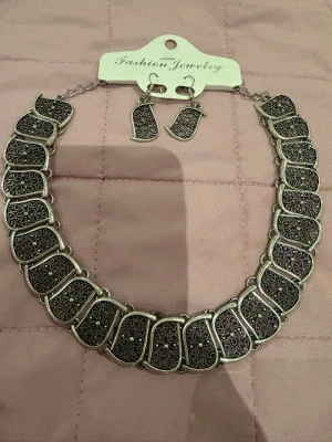 Set med halsband och örhängen i silverfärg - Säljer ett matchande set med halsband och örhängen i silverfärg. Halsbandet består av rektangulära länkar med detaljerat mönster och små glittrande stenar. Örhängena har samma mönster och form som länkarna i halsbandet. Perfekt för dig som gillar statement-smycken.