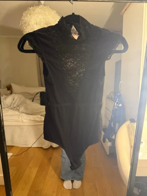 Svart spetsbody med hög krage - Säljer en svart body med hög krage och vacker spetsdetalj framtill. Bodyn har korta ärmar och en djup V-formad spetsinfällning över bröstet. Materialet är stretchigt och mjukt, perfekt för en trendig look. Ingen synlig logga eller märke.