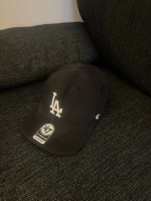 Svart LA-keps från '47 Brand - Snygg svart keps från '47 Brand med LA-logga broderad i vitt framtill. Klassisk böjd skärm och justerbar baksida. Tillverkad i bomull för en bekväm känsla. Perfekt för dig som gillar streetwear och sportig stil.