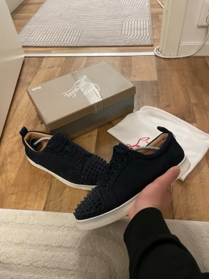 mörkblåa Christian Louboutin sneakers med nitar - Snygga svarta sneakers från Christian Louboutin i mocka med coola svarta nitar på tån och klassisk röd sula. Skorna har vita sulor och svarta skosnören, samt en låg profil som ger en stilren look. Perfekta för dig som vill sticka ut med exklusiva detaljer. Storlek 43 passar 42 obs box medföljer ej 
