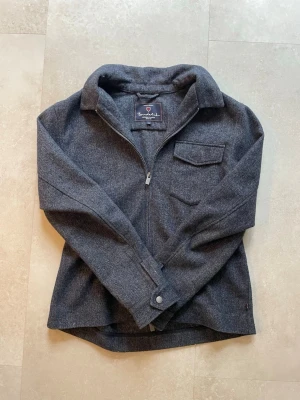 Mörkgrå Overshirt - Stilren mörkgrå overshirt med dragkedja framtill, klassisk krage och bröstficka med lock. Tillverkad i ett mjukt, ull-liknande material som ger en avslappnad men ändå snygg look. Perfekt att bära över en t-shirt.