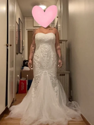 Vit axelbandslös brudklänning spets - Elegant vit brudklänning med axelbandslös design och vacker spetsdetalj över hela klänningen. Klänningen har en figurnära passform upptill och en lång, utsvängd kjol i tyll som ger en dramatisk siluett. Perfekt för dig som vill ha en klassisk och romantisk look.