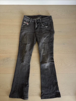 Svart/gråa bootcut jeans från Diesel - Säljer ett par svarta bootcut jeans från Diesel med slitna detaljer och klassisk femficksmodell. Jeansen har en snygg tvätt med ljusare partier och logga på myntfickan. Perfekta för dig som gillar en avslappnad, men tidlös stil.