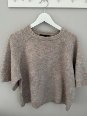 Beige stickad tröja med kort ärm - Mysig beige stickad tröja med korta ärmar och rund hals. Tröjan har en enkel, avslappnad passform och ribbade kanter vid hals, ärmslut och nederkant. Perfekt för lager på lager och passar till det mesta i garderoben.