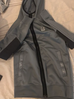 Nike Air Max grå hoodie med dragkedja - Snygg grå hoodie från Nike Air Max med hel dragkedja framtill och två fickor med Air Max-detaljer. Hoodien har en huva och svarta detaljer på ärmarna. Perfekt för en sportig och avslappnad stil. Materialet känns mjukt och har en lätt glans. Byxor ingår.