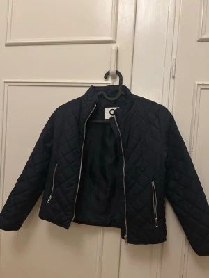 Mörkblå quiltad jacka med dragkedjor - Mörkblå svart quiltad jacka med ståkrage och två dragkedjeförsedda fickor framtill. Jackan har ett stilrent rutmönster och stängs med dragkedja. Perfekt för dig som vill ha en enkel men trendig jacka till vardagslooken.