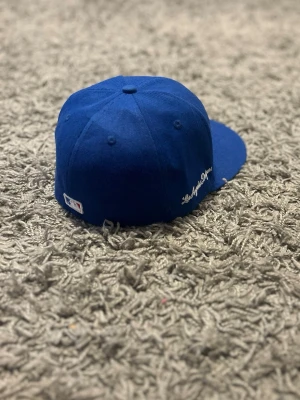 Blå LA Dodgers keps från New Era - Säljer en blå keps från New Era med Los Angeles Dodgers broderat i vitt framtill med moln. Klassisk rak skärm och snygg text på sidan. Perfekt för dig som gillar sportig streetstyle och vill ha en ikonisk accessoar.