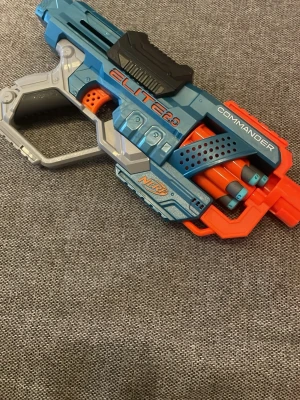 Nerf Elite 2.0 Commander Blaster - Säljer en blå och orange Nerf Elite 2.0 Commander leksakspistol. Den har plats för flera skumdartpilar och är tillverkad i plast. Perfekt för roliga Nerf-strider med vänner. Snygg futuristisk design och bekvämt handtag.