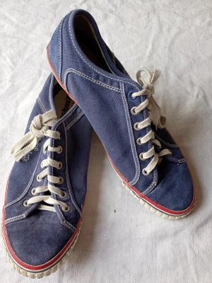 Blå sneakers från Dockers Denim - Snygga blå sneakers från Dockers Denim i canvasmaterial med vita kontrastsömmar och vita snören. Skorna har en låg profil och en vit sula med röd rand runt om. Perfekta för en avslappnad och trendig stil.