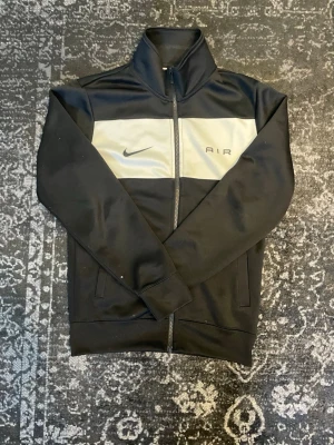 Svart Nike Air zip tröja - Svart zip tröja från Nike med en vit panel över bröstet och Nike Air-logga. Tröjan har hög krage, dragkedja hela vägen och två fickor framtill. Tillverkad i mjukt och stretchigt material som passar perfekt till en sportig stil.
