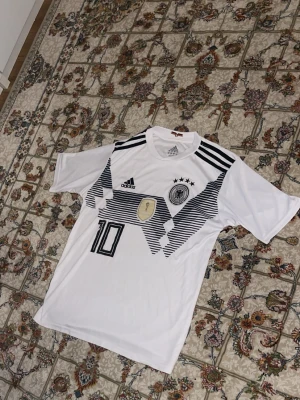 Tysklands matchtröja Adidas vit - Vit Tyskland fotbollströja från Adidas med svarta grafiska detaljer, fyra stjärnor ovanför landslagsemblemet, VM-märke på bröstet och nummer 10 tryckt framtill. Klassisk rund hals och svarta ränder på axlarna. Perfekt för fotbollsfans!