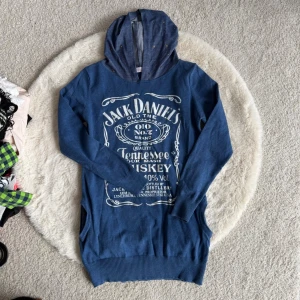 Blå Jack Daniel's hoodie-tröja - Blå hoodie med vit Jack Daniel's-tryck framtill och lång ärm. Huvan har små nitar som detalj och insidan är mönstrad. Tröjan har ribbade muddar vid ärmslut och nederkant. Perfekt för dig som gillar streetstyle och vill sticka ut.