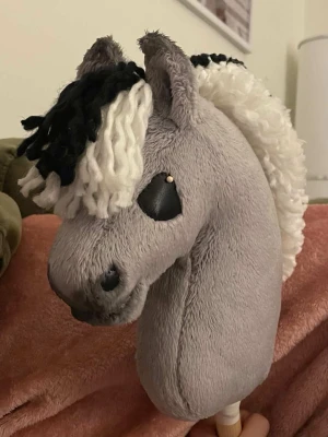Grå käpphäst med svartvit man - Säljer en grå käpphäst med mjuk plysch, svart och vit garnman samt svarta tygögon. Hästen har detaljerade öron och nos.  Perfekt för lek och hobbyridning!