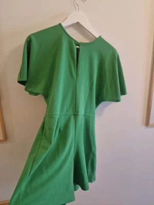 Grön kortärmad jumpsuit - Snygg grön jumpsuit med korta vida ärmar och rund halsringning. Modellen har en markerad midja och vida shorts. Perfekt för dig som vill ha en bekväm och trendig look. Materialet känns mjukt och stretchigt. Några tråddragningar på framsidan.