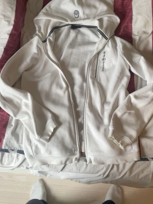 Vit hoodie från Sail Racing - Säljer en vit hoodie från Sail Racing med dragkedja framtill och broderad logga på bröstet. Hoodien har en klassisk huva och ribbade muddar vid ärmslut och nederkant. Tillverkad i mjukt bomullsmaterial som är skönt mot huden. Perfekt för en avslappnad och sportig stil.