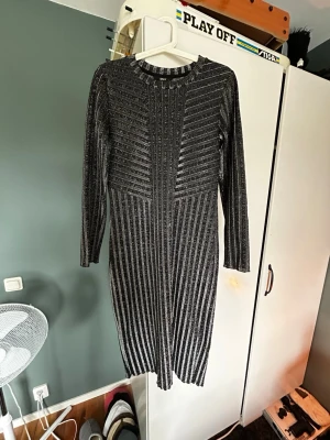 Glittrig svart långklänning - Snygg svart långklänning med glittriga silverränder och lång ärm. Klänningen har en kroppsnära passform och rund halsringning. Perfekt för dig som vill sticka ut med en elegant look och gillar metallicdetaljer.