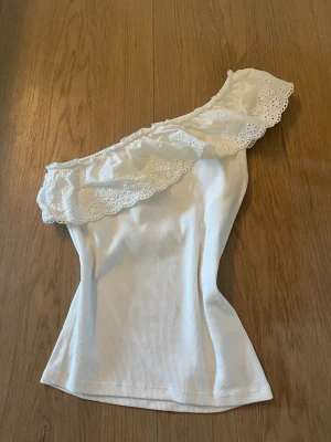 Vit volang topp - Snygg vit oneshoulder topp från Kappahl med ribbad struktur och bred volangkant i spets längs axeln. Perfekt för dig som vill ha en unik och trendig look. Materialet känns mjukt och följsamt. Stoelk 170 (ca xs)