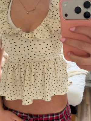 Prickig volangtopp med knyt i ryggen - Supersöt topp i ljus beige med svarta prickar, volangkant vid urringningen och knytband i ryggen. Modellen är ärmlös och har en lätt utsvängd passform. Perfekt för dig som gillar en lekfull och trendig stil.