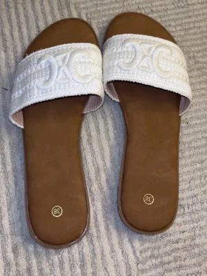 Vita sandaler med broderad logga - Vita slip-in sandaler med broderad detalj framtill och brun sula. Platt modell som är bekväm och enkel att bära. Perfekta för sommar, semester eller vardag. Oanvända, sulorna i fint skick.🖤