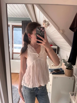 Vit volangblus med v-ringning - Supersöt vit blus med volangdetaljer och v-ringning. Blusen är ärmlös och har en luftig, flowy passform som känns riktigt fräsch till jeans eller kjol. Perfekt för varmare dagar och ger en romantisk vibe till din outfit. Kan bäras åt båda håll.