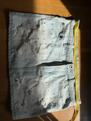 Ljusblå jeanskjol med slitningar och nitar - Trendig ljusblå jeanskjol med slitna detaljer och små glittriga nitar över hela framsidan. Klassisk femficksmodell med bälteshällor och knappstängning. Perfekt för dig som gillar en edgy och cool stil.