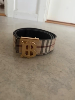 Burberry rutigt bälte med guldspänne - Snyggt bälte från Burberry med klassiskt rutigt mönster i beige, brunt och rött. Bältet har ett stort guldigt spänne med Burberrys logotyp och är tillverkat i skinn. Perfekt accessoar för att lyfta din outfit.
