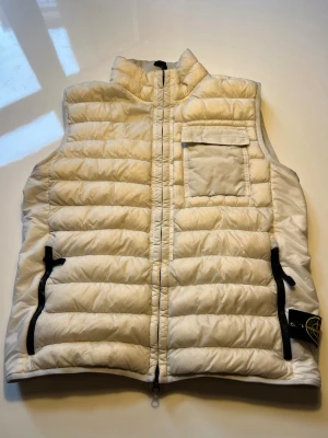 Vit dunväst från Stone Island - Dunväst från Stone Island med quiltad design och hög krage. Västen har en bröstficka med lock, två svarta dragkedjefickor och klassisk Stone Island-patch på sidan.