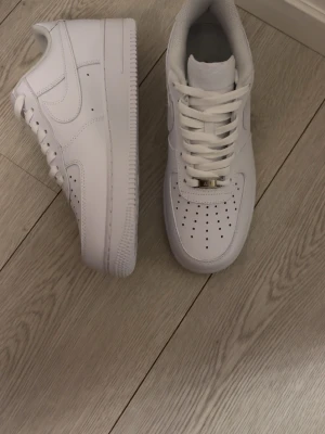 Nike Air Force  - Klassiska Nike Air Force 1 sneakers i helvitt läder med perforerad tåbox, vit sula och matchande vita skosnören. Ikonisk design med Nike swoosh på sidorna och metallbricka på snörningen. Perfekta för en clean och stilren look.