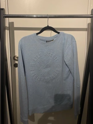 Ljusblå sweatshirt från Lyle & Scott - Ljusblå sweatshirt med rund hals från Lyle & Scott. Tröjan har ett stort broderat logotyptryck framtill med text och en fågel. Mjuk bomullskänsla, ribbade muddar vid ärmslut och nederkant. Perfekt för en chill och stilren look.