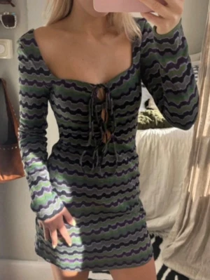 Mönstrad kortklänning missoni vibe - Säljer en vågig mönstrad kortklänning från Zara i grönt, svart och lila. Klänningen har lång ärm, snörning framtill och en figurnära passform. Perfekt för dig som gillar unika och färgglada plagg.