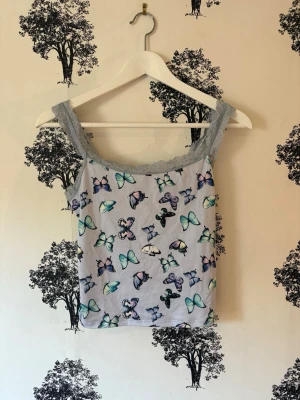 Ljublå linnetopp med fjärilar - Söt ljusblå linnetopp med spetskant och fjärilsmönster i flera färger. Toppen har breda axelband i spets och croppad passform. Perfekt för dig som gillar gulliga detaljer och en lekfull stil!🦋💓🌸