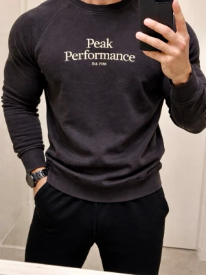 Svart sweatshirt från Peak Performance - Klassisk svart sweatshirt från Peak Performance med vit logga på bröstet. Tröjan har rund halsringning, långa ärmar och ribbade muddar vid ärmslut och nederkant. Perfekt för en avslappnad och stilren look.