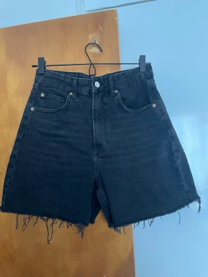 Svarta högmidjade jeansshorts - Svarta högmidjade jeansshorts från Pull&Bear med råa, fransiga benslut och klassisk femficksdesign. Shortsen har en knapp och dragkedja framtill samt bälteshällor. Perfekta för en avslappnad och trendig look.