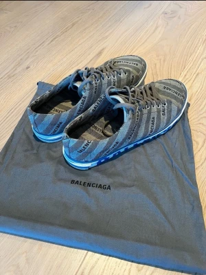 Grå Balenciaga sneakers med logga - Snygga grå sneakers från Balenciaga med tydlig logotyp i svart över hela ovandelen och stor vit/blå logga på sulan. Skorna har lågt skaft, klassisk snörning och är tillverkade i textilmaterial. Perfekta för dig som gillar streetwear och vill sticka ut. Skriv för mer info. Storlek 42 och är autentisk