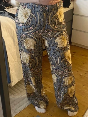 Vida kostymbyxor med blommönster - Säljer ett par riktigt snygga kostymbyxor med blommigt mönster i beige, grått och inslag av rosa. Byxorna har hög/mid midja och vida ben för en avslappnad men trendig look. Perfekta för dig som vill sticka ut med din stil. Materialet känns mjukt och följsamt. Säljer för lite mer eftersom det är ett så pass känt mönster och inom en kollektion som tog slut direkt samt inte finns så många av. Super fina och eleganta, kan självklart diskutera pris med mera, finns bundle rabatter! 