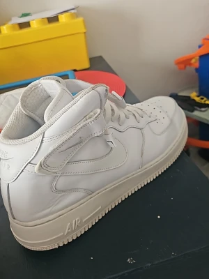 Nike Air Force 1 vita high sneakers - Klassiska Nike Air Force 1 high sneakers i helvitt läder med vit sula och snörning. Skorna har en hög siluett med kardborreband runt ankeln och ikonisk Swoosh-logga på sidan. Perfekta för en clean och stilren streetlook.