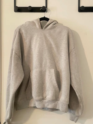 Ljusgrå hoodie med magficka - En stilren ljusgrå hoodie med huva och stor magficka framtill. Tröjan har ribbade muddar vid ärmslut och nederkant, samt en avslappnad passform. Perfekt för en chill och avslappnad look. Materialet känns mjukt och bekvämt mot huden.