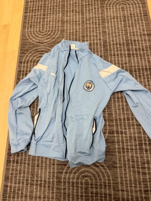 Manchester City ljusblå träningsjacka - Ljusblå träningsjacka från Puma med Manchester Citys klubbmärke på bröstet. Jackan har vita detaljer på axlarna, svarta dragkedjor och ståkrage. Perfekt för fotbollsträning eller att bära till och från gymmet.