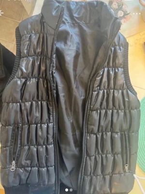 Svart väst - Snygg svart dunväst med glansig finish och quiltad design. Västen har hög krage, två fickor med dragkedja och ribbade kanter vid ärmhålen. Perfekt för lager på lager och ger en cool streetwear-look.
