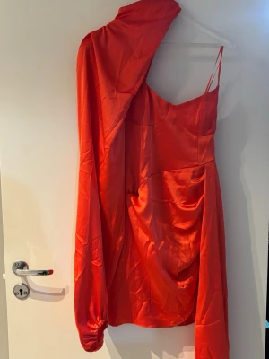 Röd oneshoulder klänning i satin - Cool röd klänning med oneshoulder-design som är en lång ärm. Klänningen har en asymmetrisk form och är gjord i glansigt satinmaterial som ger en lyxig känsla. Perfekt för dig som vill sticka ut och ha något unikt på nästa event.
