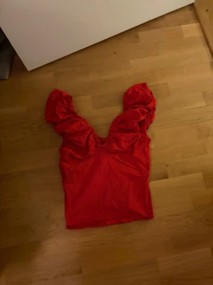 Röd volangblus från Holly Whyte - Snygg röd blus från Holly Whyte med volangdetaljer på axlarna och djup v-ringning. Toppen har en feminin och trendig look, perfekt för att sticka ut. Tillverkad i mjuk bomull som känns skön mot huden.