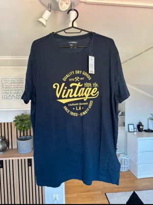 Mörkblå Vintage T-shirt från Livergy - Snygg mörkblå T-shirt från Livergy med gul vintage-tryck framtill. Klassisk rund halsringning och korta ärmar. Tillverkad i mjuk bomull som är skön att bära. Perfekt för en avslappnad och trendig look.