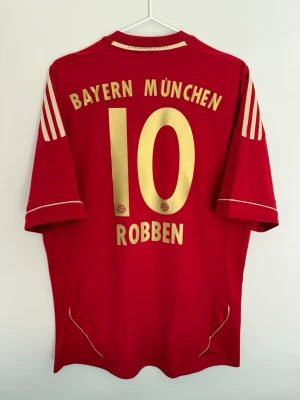 Bayern München 11/12 Home - #10 Robben - size L - 8/10 - Bayern Münchens home kit in red and gold used during both 2011/12 and 2012/13 seasons. On the back, the class winger Arjen Robben with number 10.  Condition: 8/10. Some cracks at the front sponsor. The Bayern Munchen text at the back has been remade which can be seen from closer range.   Size: L Produktkod: V13554